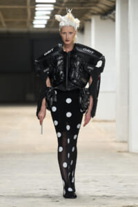 Model wearing futuristic black protective armor labeled GRIT over a black polka-dot dress on a runway with stark white hair.
