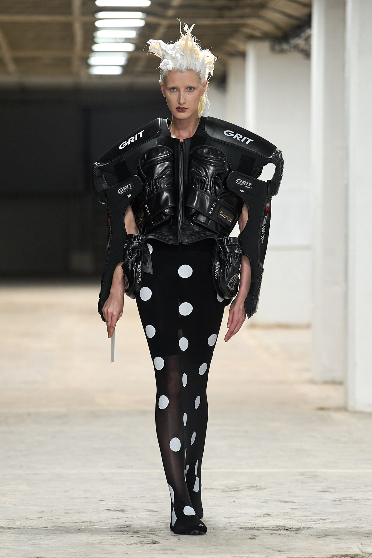 Model wearing futuristic black protective armor labeled GRIT over a black polka-dot dress on a runway with stark white hair.
