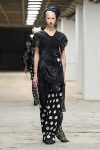 Model walks the runway in a black lace and satin dress paired with black-and-white polka-dot pants, wearing a black headwrap.