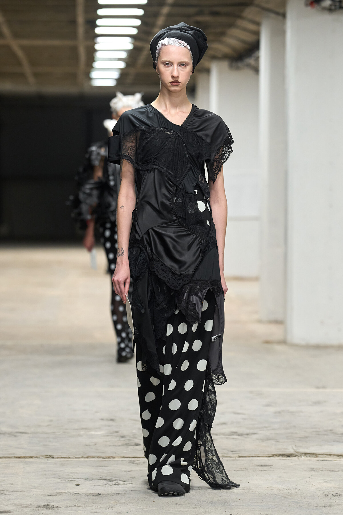 Model walks the runway in a black lace and satin dress paired with black-and-white polka-dot pants, wearing a black headwrap.