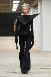 Model in an all-black avant-garde gown with a large angular collar walking a runway.