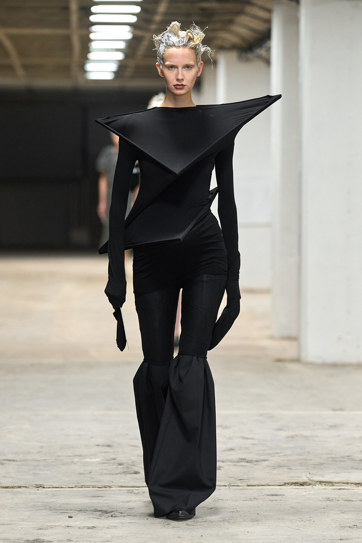 Model in an all-black avant-garde gown with a large angular collar walking a runway.
