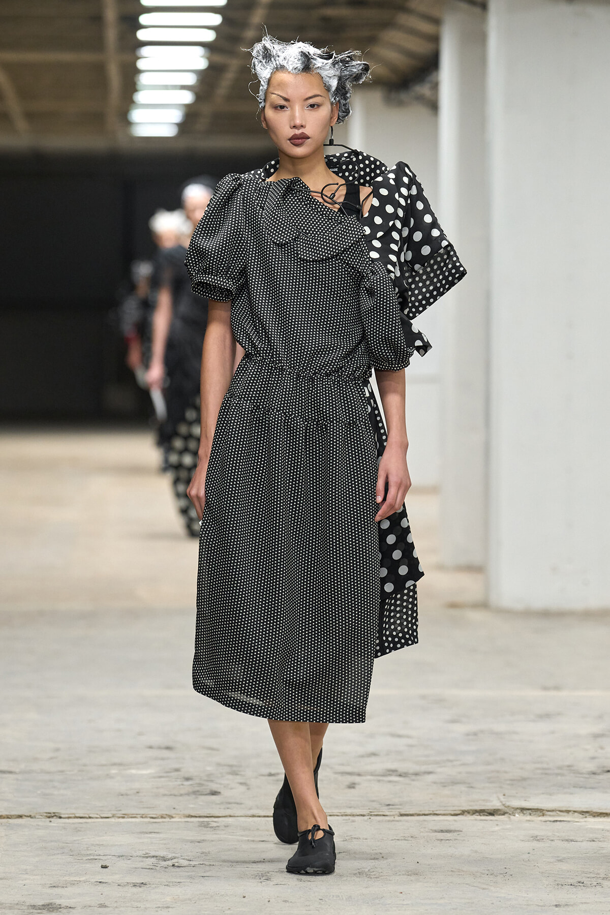 Model walking a runway in a black-and-white polka-dot dress with puff sleeves and a draped shoulder detail, wearing black flats and a metallic hair accessory.
