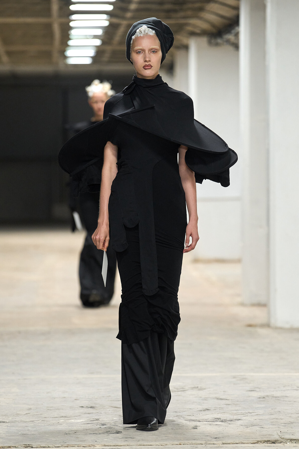 Model walking the runway in an all-black avant-garde ensemble with large, sculptural shoulder pieces and a head wrap.