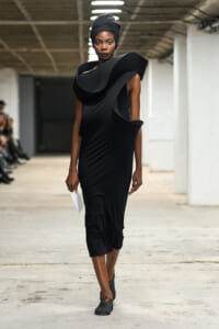 Model walking a minimalist black sculptural dress with bold shoulder pieces and a black headwrap in an industrial runway setting; holds a white envelope in her left hand.