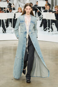 Model walking a runway in a light denim long coat over a sheer lace blouse and black pants, with an orchestra in the background.