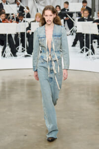 Model walks the runway in a light denim jumpsuit with tied waist sash, deep neckline, and wide legs, backstage orchestra in the background.