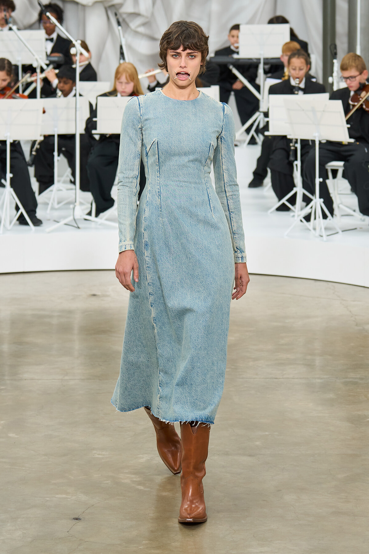 Model walks the runway in a light-blue denim dress with a frayed hem and brown boots, orchestra musicians visible in the background.] ,