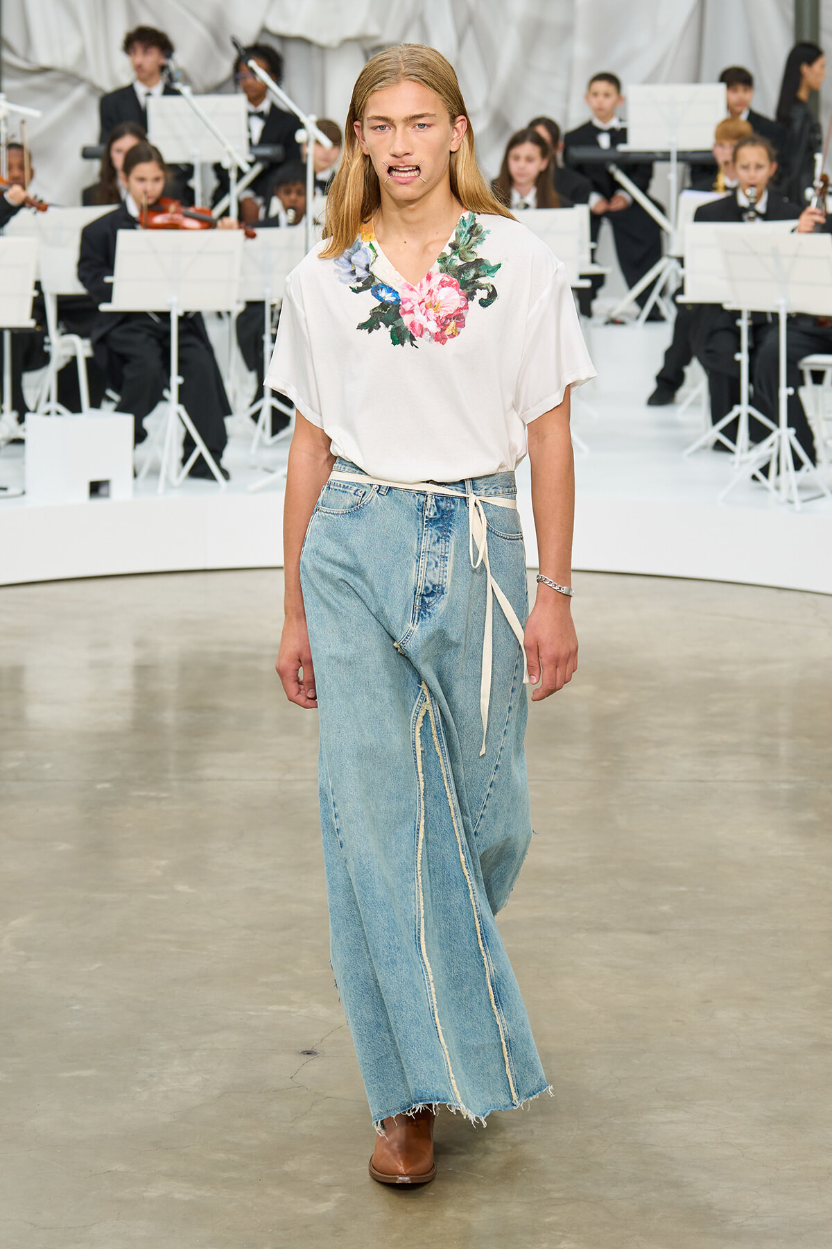 Model walking a fashion runway in a white floral top and wide-leg light denim jeans with a wrap belt, orchestra visible in the background.