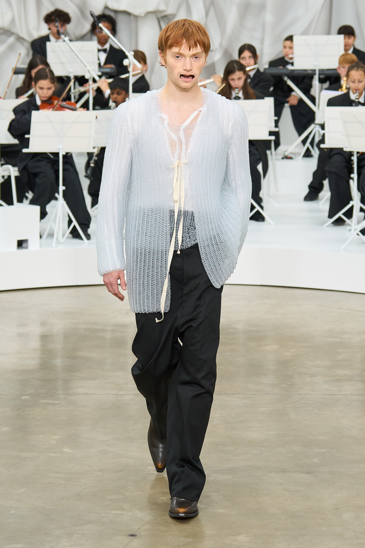 Male model walks the runway wearing a sheer, light-knit cardigan over an open white blouse, paired with black trousers and brown shoes, backstage orchestra in the background