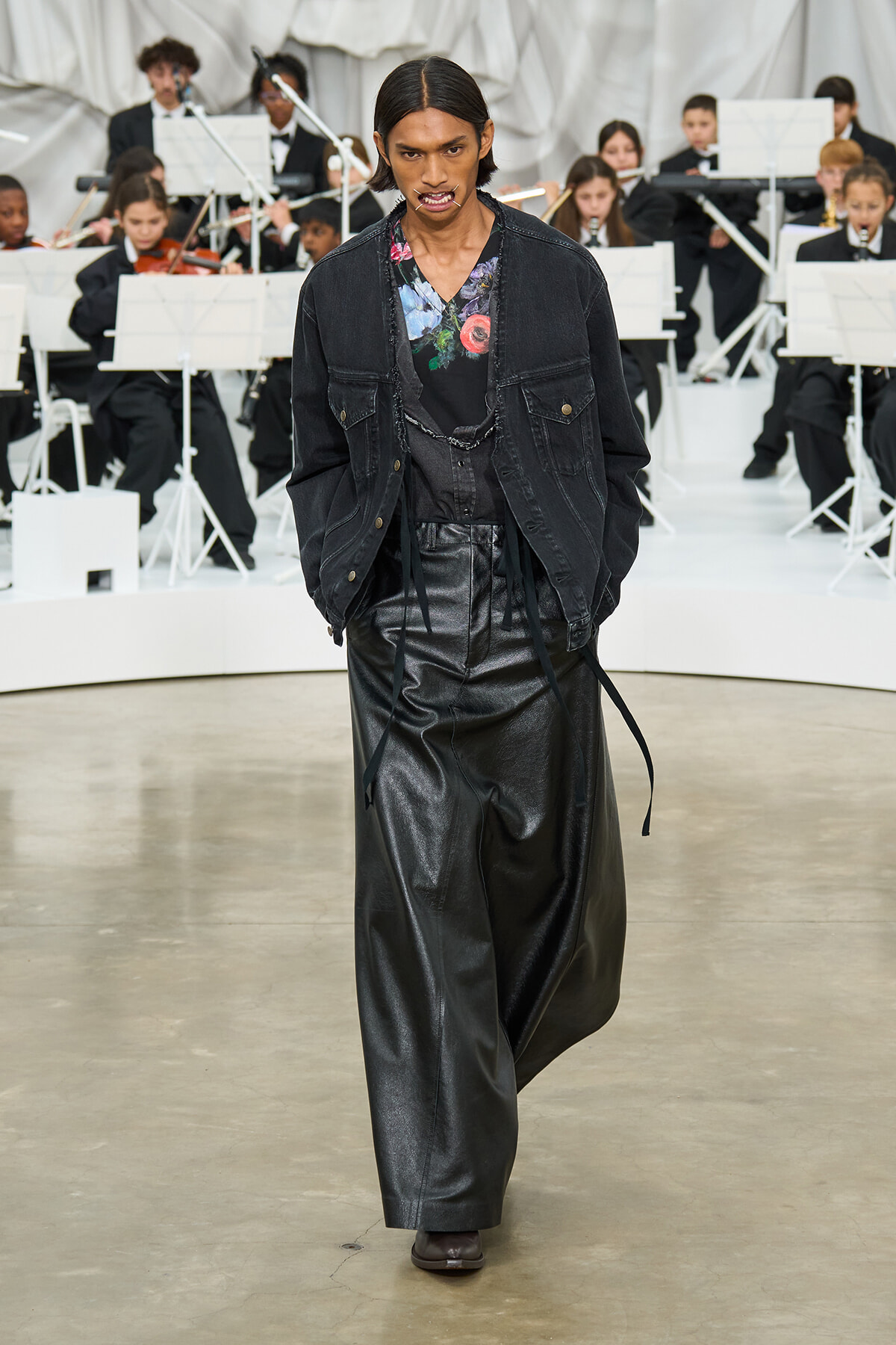 Model walks the runway in a black denim jacket over a floral top and wide-leg black leather pants; an orchestra sits in the background.