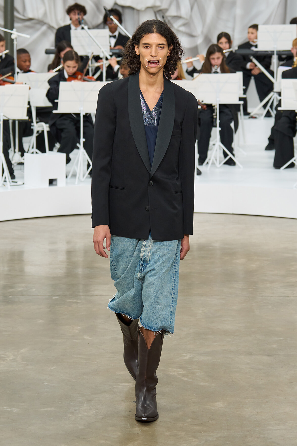 Male model walking a runway in a black satin-lapel blazer, dark top, oversized frayed denim shorts, and tall brown boots, with an orchestra in the background.
