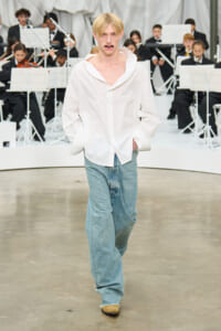 Model walking a runway in an oversized white shirt and baggy light-blue jeans, with a seated orchestra in the background.