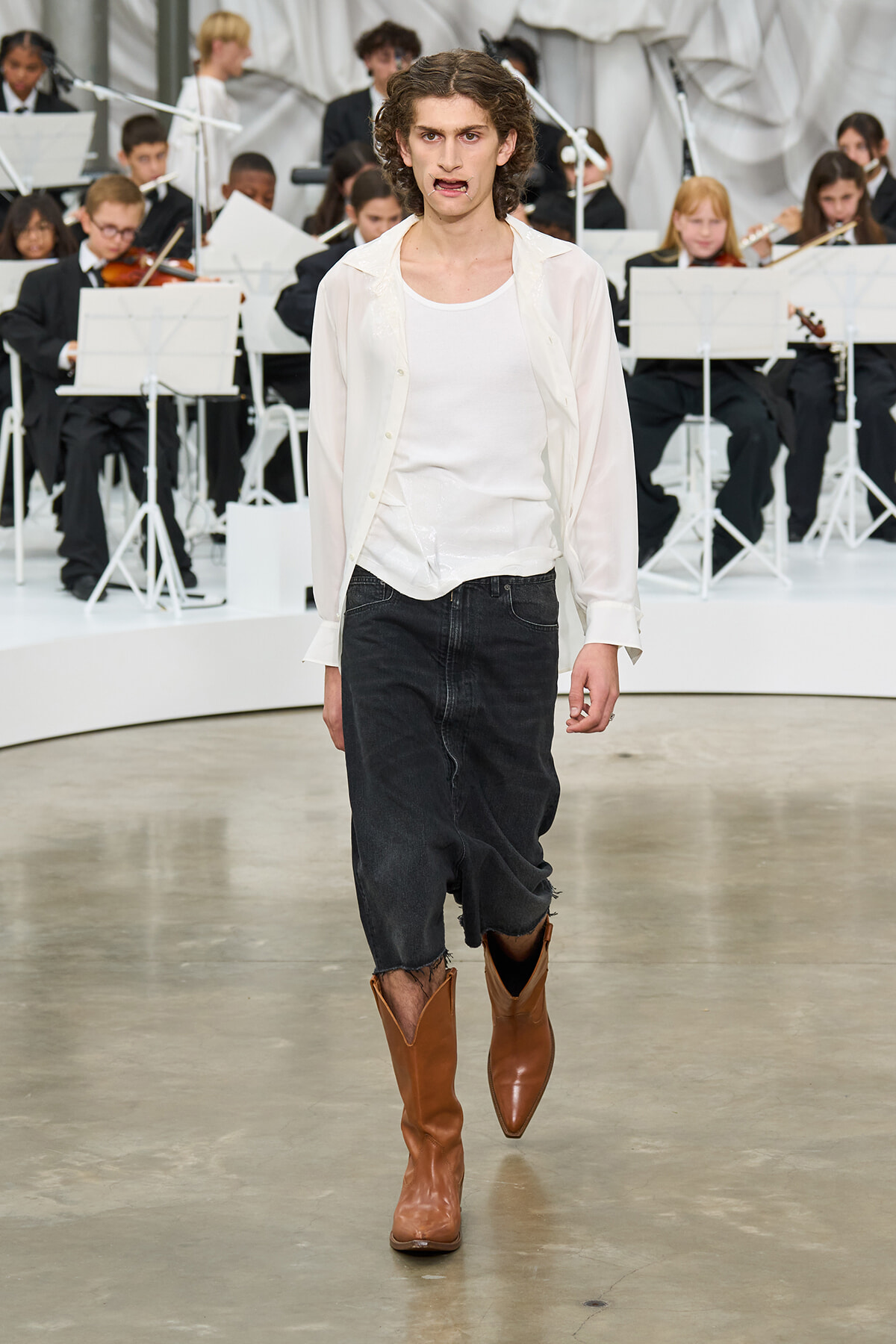 Model walking the runway in a white sheer shirt, black denim, and brown cowboy boots, with an orchestra in the background.