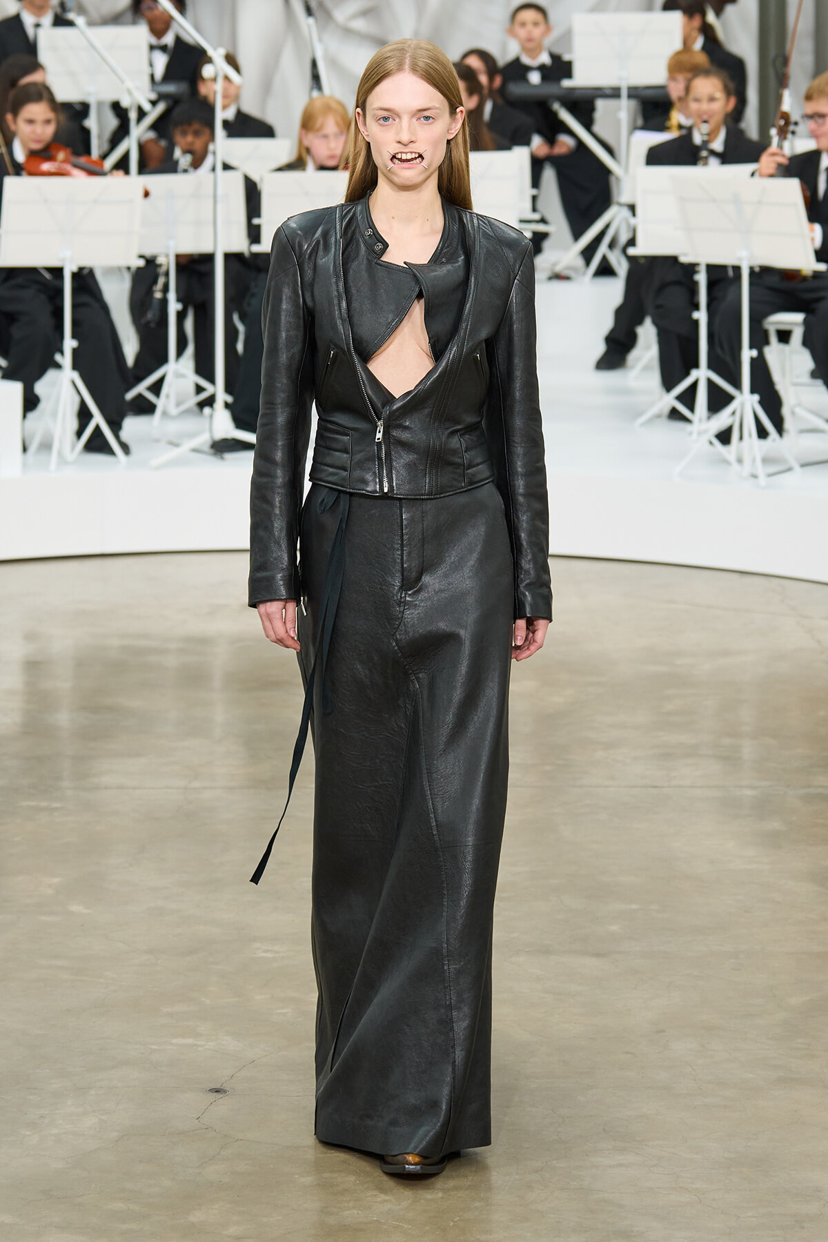 Model walking the runway in a black leather two-piece outfit with a cutout top, long skirt, and belt detailing, with an orchestra in the background.