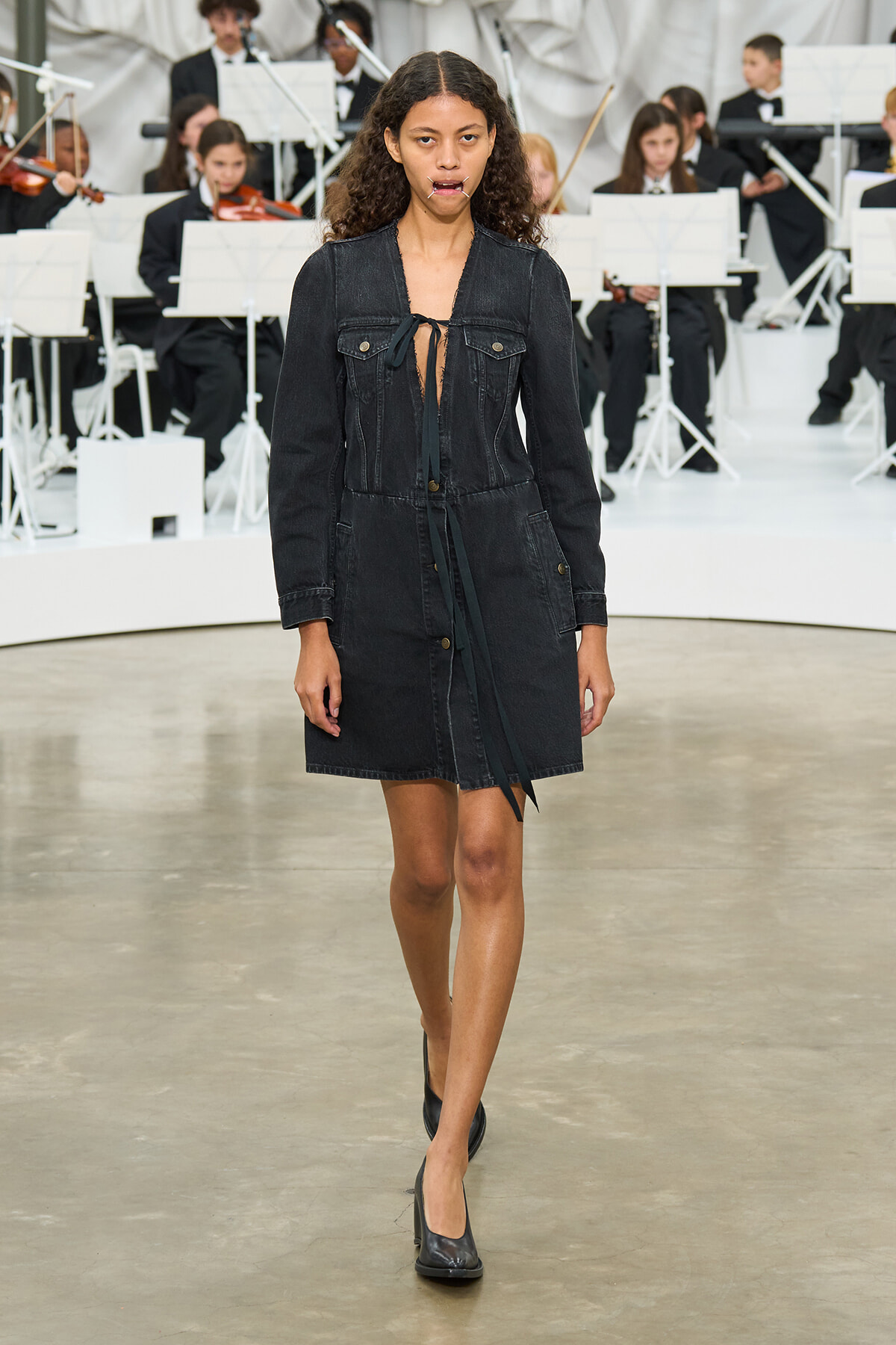 Model walks a runway in a black denim shirt-dress with a tie at the neckline; wearing black heels, with an orchestra in the background.