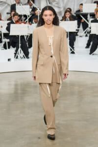 Models wears a beige tailored suit with a long blazer and wide trousers on a runway, an orchestra backdrop behind her