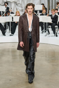Male model on a runway wearing a long brown leather coat over a white shirt and shiny black pants, with an orchestra in the background.
