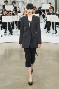Model walking a fashion runway in a dark oversized blazer, black cropped pants, and black heels with a beige top and tie detail, an orchestra visible in the background