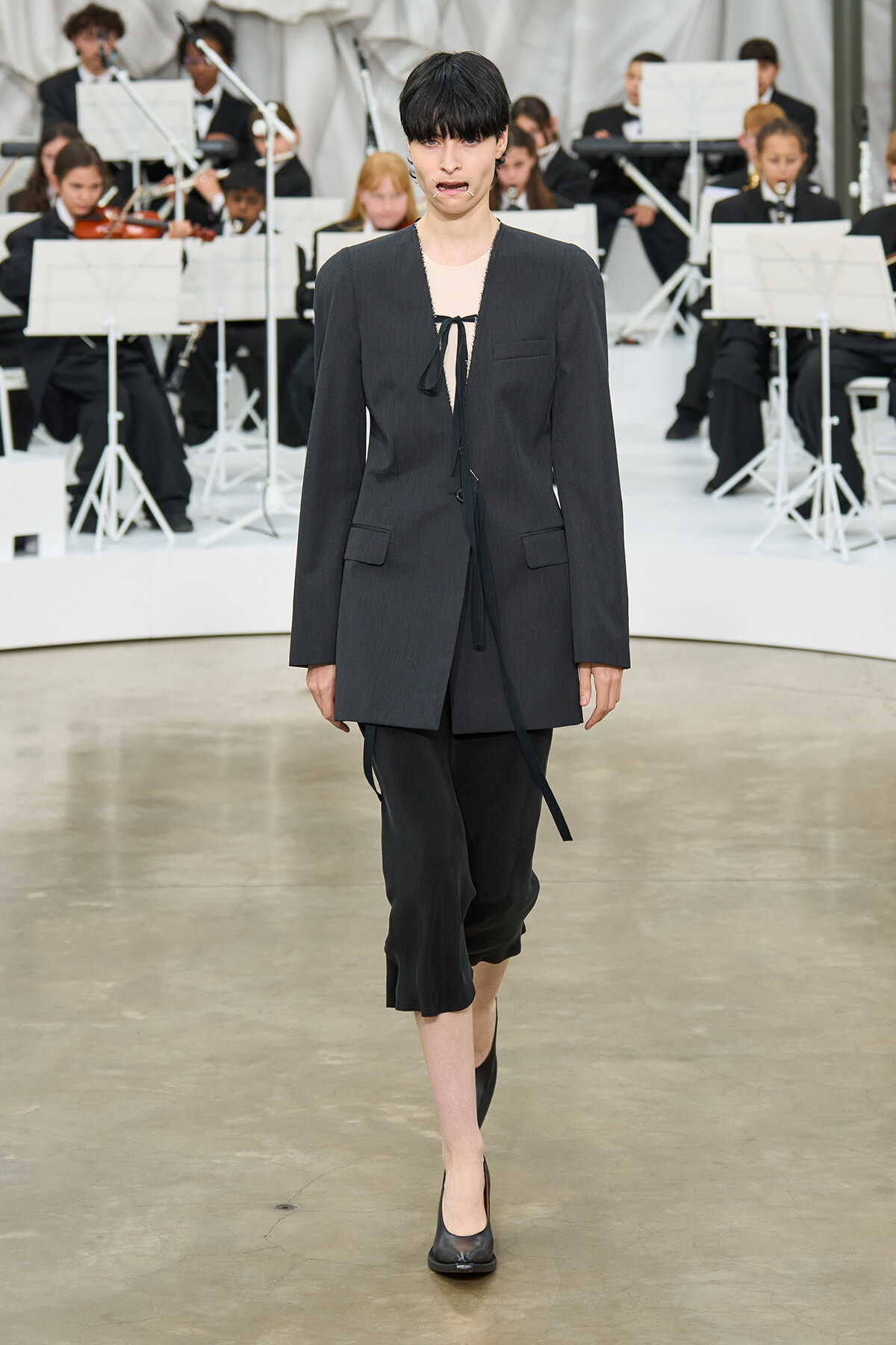 Model walking a fashion runway in a dark oversized blazer, black cropped pants, and black heels with a beige top and tie detail, an orchestra visible in the background