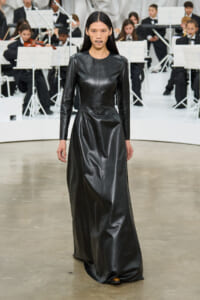 Model walking the runway in a long black leather dress, in front of an orchestra on stage.