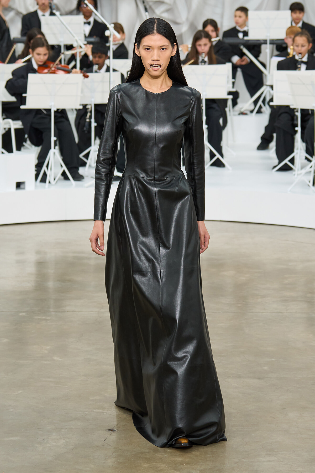 Model walking the runway in a long black leather dress, in front of an orchestra on stage.