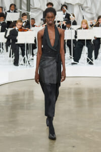 Model walking a runway in a black halter dress with an overlapping leather apron, paired with black boots, as a classical orchestra performs in the background.