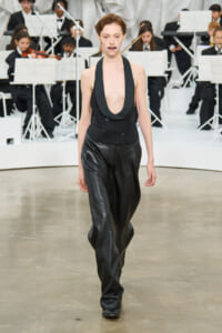 Model walking a fashion show runway in a black deep-plunge halter blazer and black leather pants with an orchestra in the background