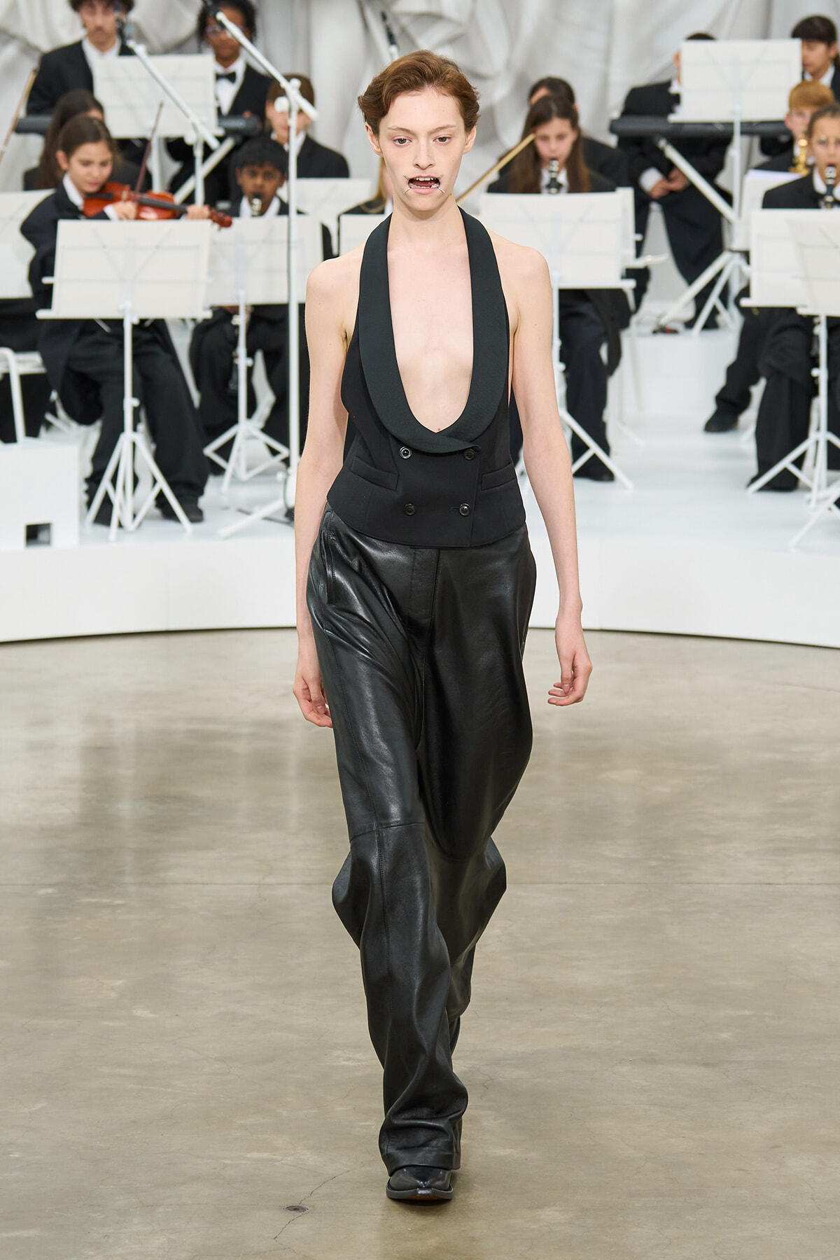 Model walking a fashion show runway in a black deep-plunge halter blazer and black leather pants with an orchestra in the background