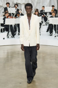 Male model on runway wearing a cream blazer, white shirt, and dark jeans with a band in the background.