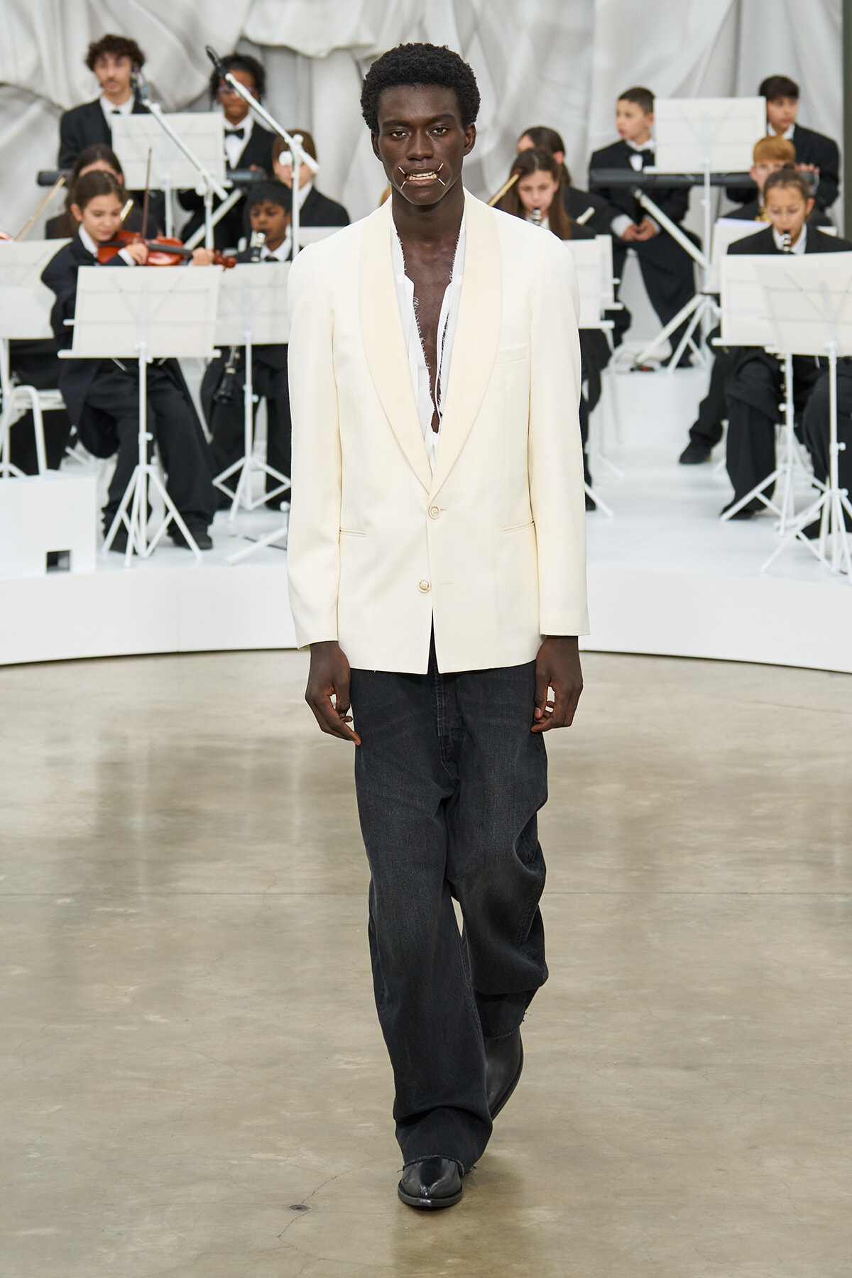 Male model on runway wearing a cream blazer, white shirt, and dark jeans with a band in the background.