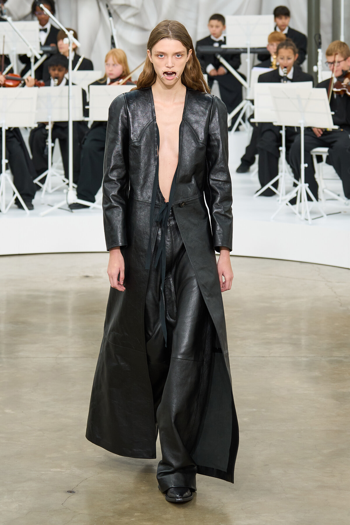 Model walking the runway in a long black leather coat opened at the front with a belt, paired with black pants; a full orchestra is visible in the background