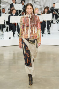 Model walking a runway wearing a metallic, multicolored textured jacket and a layered, patterned skirt with boots, on a white stage with an orchestra in the background.