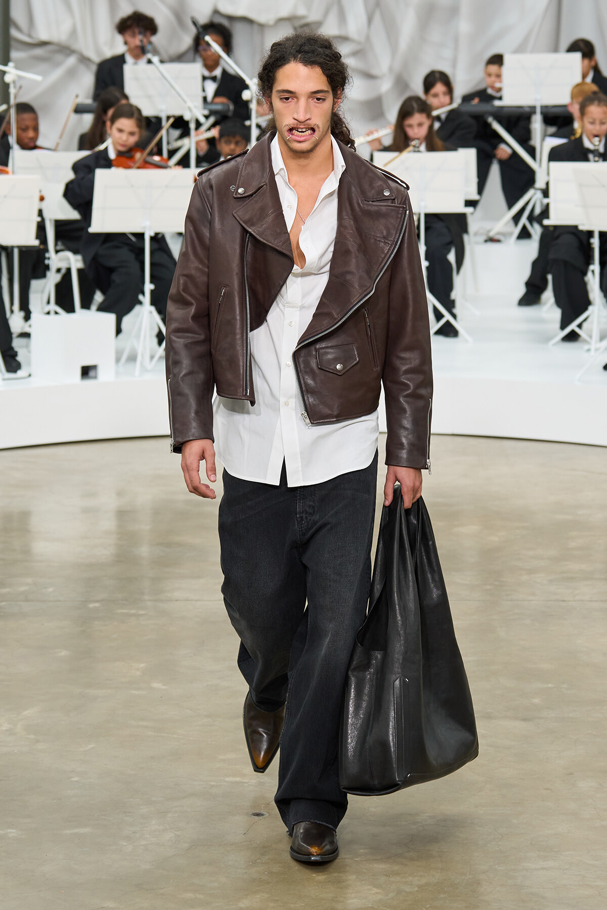 Male model walking on runway in a brown leather jacket, white shirt, dark jeans, and brown boots, carrying a large black bag with an orchestra in the background