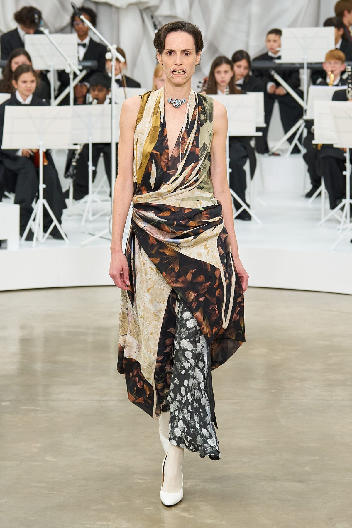 Runway model wearing a sleeveless, draped, multi-pattern gown in brown, beige and floral panels, with white ankle boots.