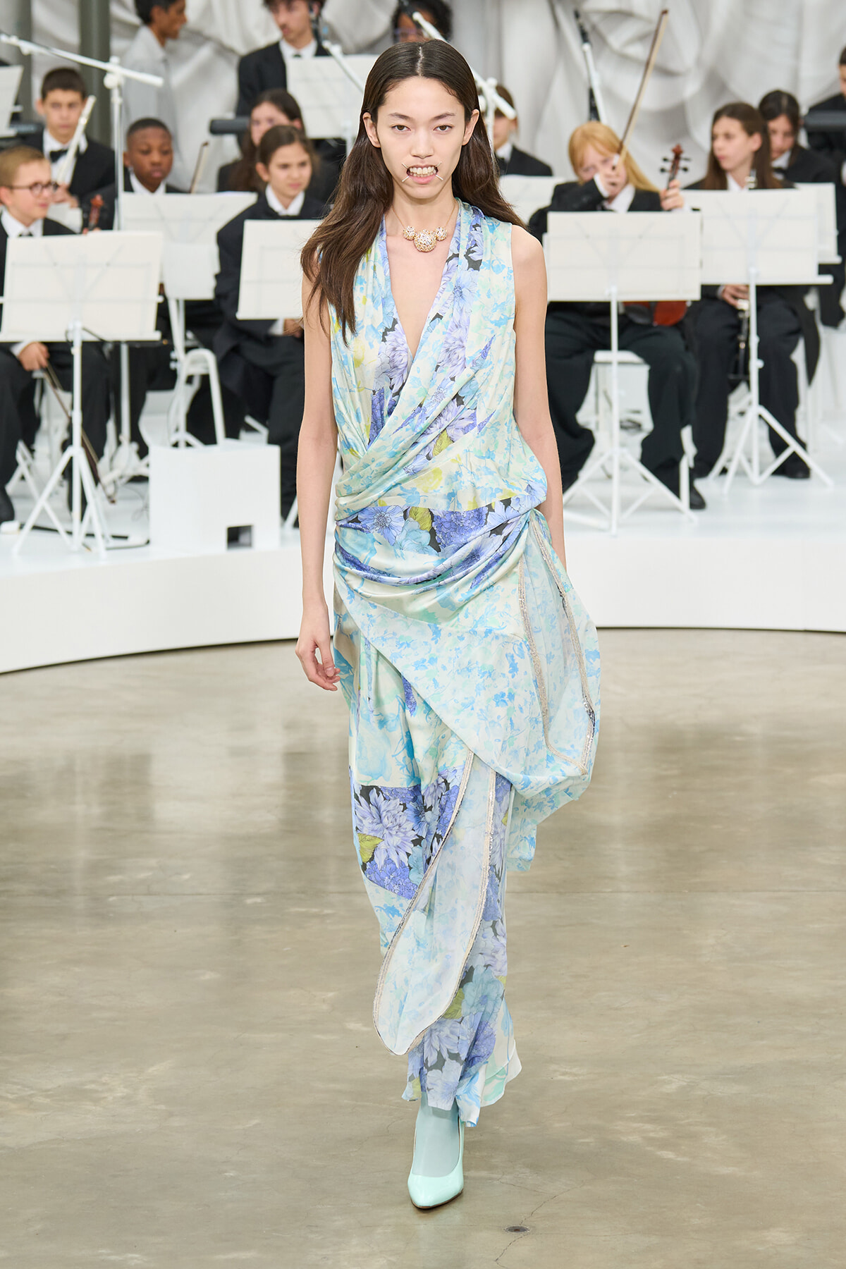 Model walking a runway in a pastel blue floral draped gown with turquoise heels, an orchestra visible in the background.