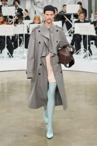 Model walking the runway in a long gray trench coat, pastel pink top, and mint tights, carrying a brown handbag with an orchestra in the background