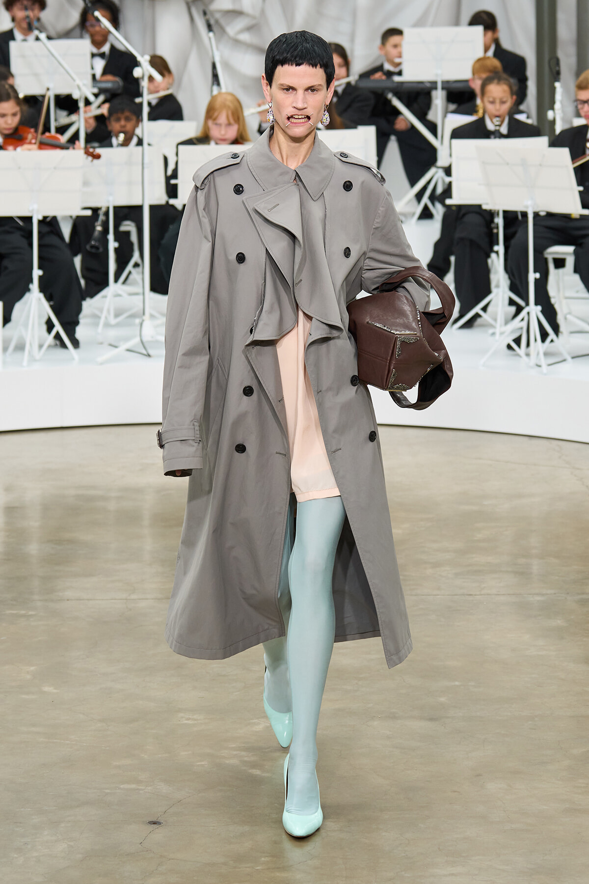 Model walking the runway in a long gray trench coat, pastel pink top, and mint tights, carrying a brown handbag with an orchestra in the background