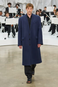 Male model walks a runway in a navy long coat with a black outfit, orchestra visible behind him on stage.