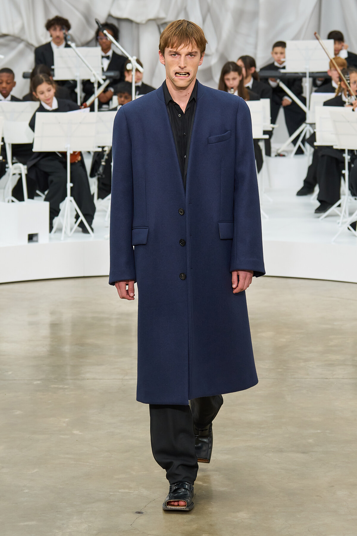 Male model walks a runway in a navy long coat with a black outfit, orchestra visible behind him on stage.