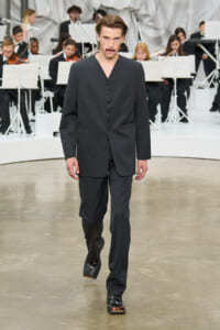 Male model walks the runway in a dark gray suit with an orchestra visible in the background.