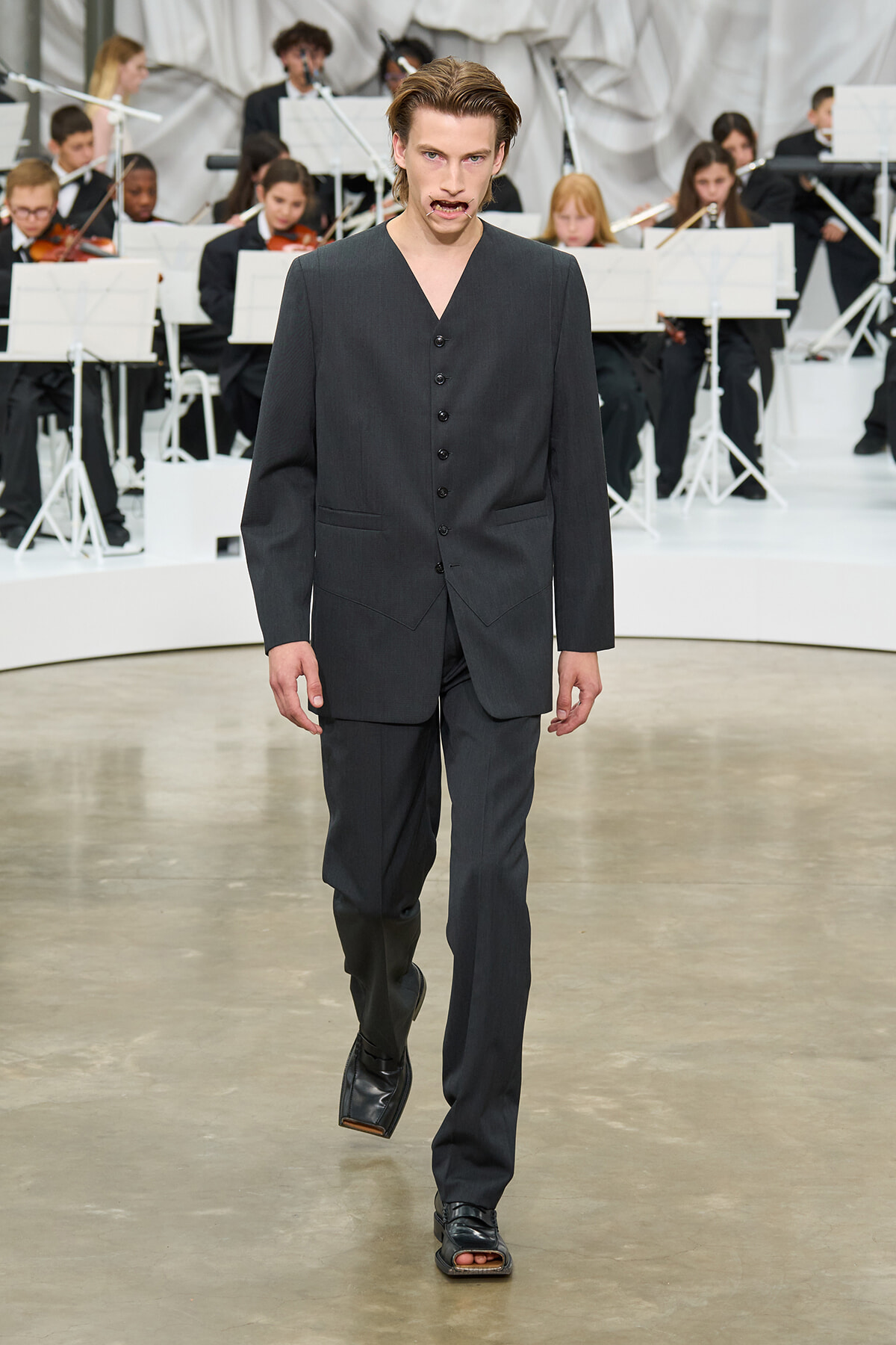 Male model walks the runway in a dark gray suit with an orchestra visible in the background.
