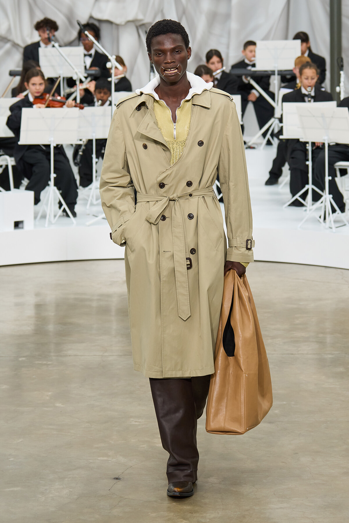 Model walking a runway in a beige trench coat, belt tied, carrying a tan leather bag with an orchestra in the background.