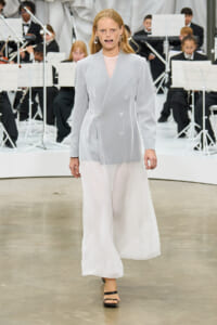 Model walks the runway in a pale gray double-breasted blazer and sheer white wide-leg pants, with an orchestra in the background.