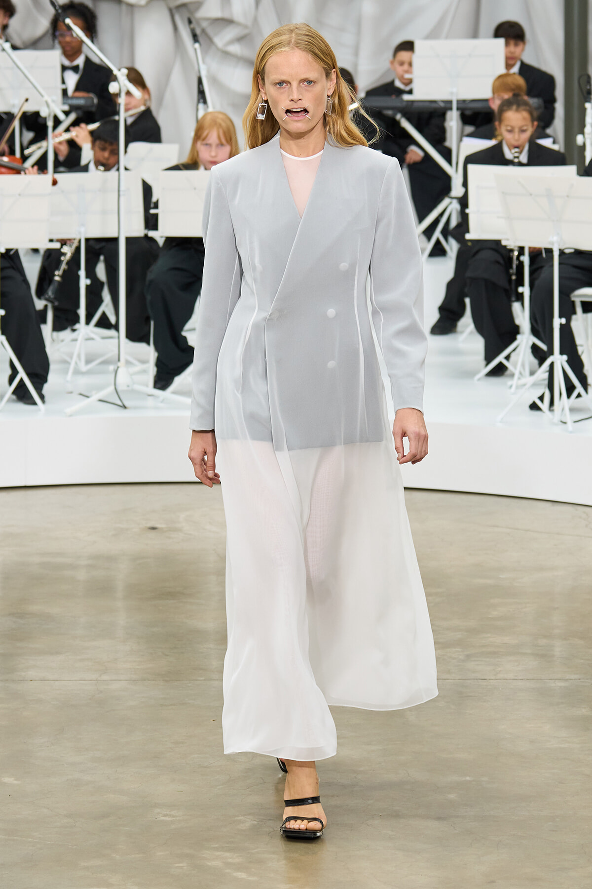 Model walks the runway in a pale gray double-breasted blazer and sheer white wide-leg pants, with an orchestra in the background.