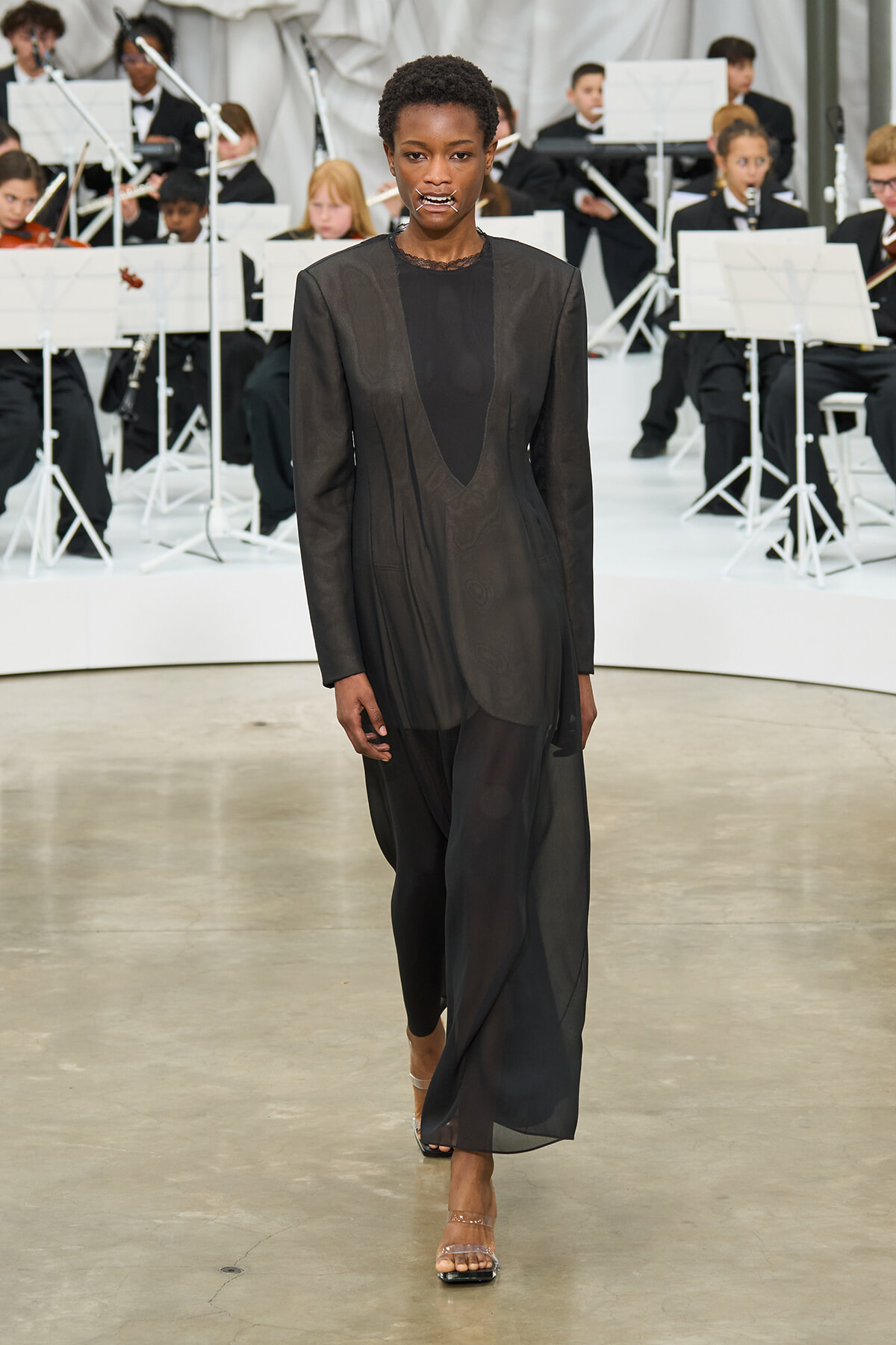 Model walks the runway in a sheer black layered suit as a live orchestra performs behind on a white stage.