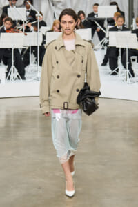 Model walks a runway in a beige double-breasted trench over pastel layered dress with lace hem, white heels, and a black clutch; seated orchestra in the background.