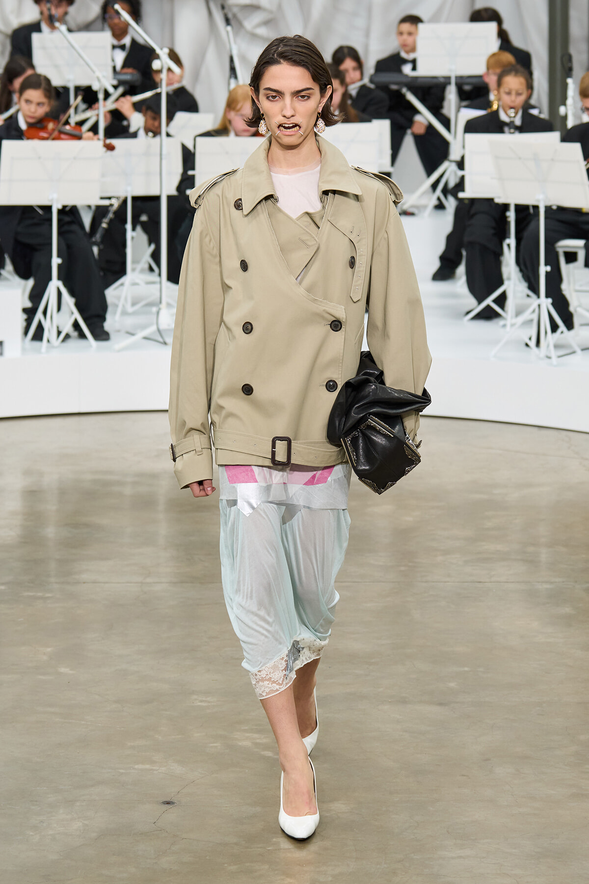 Model walks a runway in a beige double-breasted trench over pastel layered dress with lace hem, white heels, and a black clutch; seated orchestra in the background.