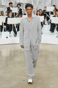 Male model walks a runway in a light gray oversized suit with an open collar, orchestra in the background.