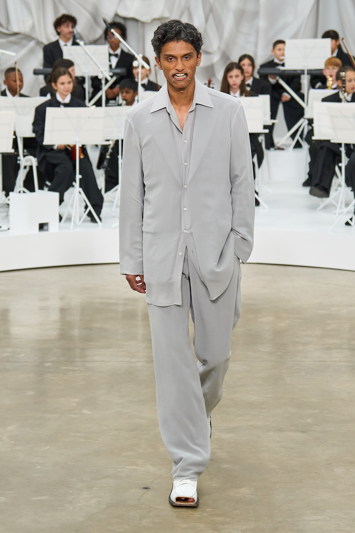 Male model walks a runway in a light gray oversized suit with an open collar, orchestra in the background.